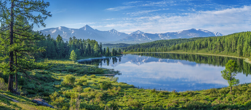 View of a mountain lake on a summer morning, picturesque reflection of the sky with clouds, snow-capped peaks and forested shores