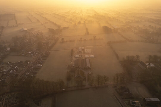 Aerial view of a farm surrounded by misty fields and trees during a golden sunrise in Friesland, Netherlands.