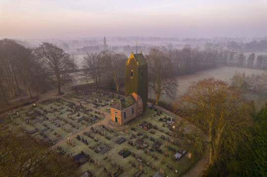 Aerial view of the historic Groene toren tower covered in ivy, standing amidst a cemetery with rows of gravestones, surrounded by trees and misty fields in Eastermar, Friesland, Netherlands.