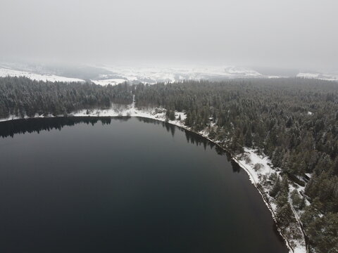 Aerial view of Lac de Serviere surrounded by snow-covered pine forests under a cloudy sky in Orcival, Auvergne-Rhone-Alpes, France.