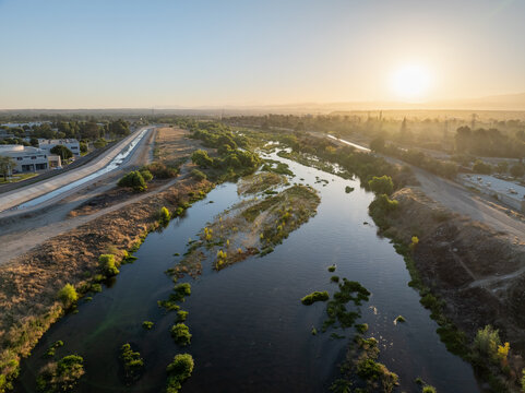 Aerial view of the Kern River with lush vegetation along the banks, dry terrain, and a concrete canal nearby under the warm light of the setting sun in Bakersfield, California, United States.