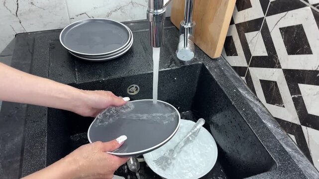 Woman rinsing a soapy plate under running water and stacking it with clean dishes in the kitchen, household routine, hygiene and domestic care.
