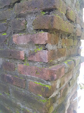 Close up of ancient weathered red brick wall corner with green moss and rough concrete texture.