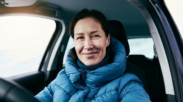 Smiling woman sitting inside a car during winter, wearing warm blue puffer jacket, enjoying travel and journey