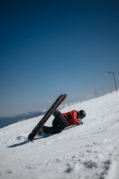 A snowboarder in a red jacket lies after a fall on a snowy ski slope. The rider wearing a helmet and goggles rests on powder snow beneath a clear blue sky.