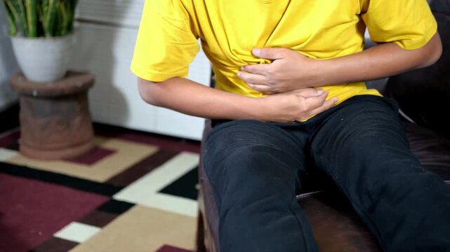 Stomach ache, stomach cramps, heartburn, digestive problems. A young Asian man is holding his stomach and grimacing in pain while sitting on the couch at home.
