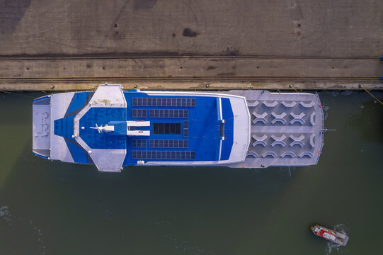 Aerial view of a ferry boat docked at a pier with solar panels on its roof and seating areas, accompanied by a small safety training life raft in the water in the Netherlands.