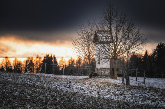 Small historical wayside shrine on a snowy field during dramatic winter sunset. Bare trees next to a traditional Czech religious column shrine with dark stormy sky and orange horizon.
