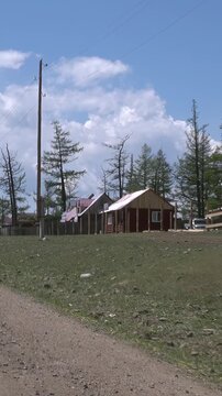 Witness a peaceful summer day by Khuvsgul Lake, Mongolia. Rustic homes stand nestled among tall trees, a dirt path winding through green fields under a vast azure sky with soft clouds.