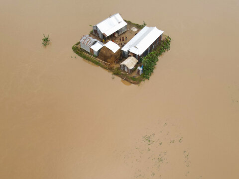 Aerial view of Sunamganj Haor showing an isolated homestead surrounded by floodwaters with corrugated metal roofs and people standing outside in Sunamganj District, Sylhet Division, Bangladesh.