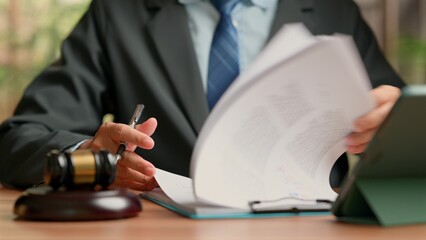 Professional male attorney wearing dark suit reviews legal document paperwork holding pen near...