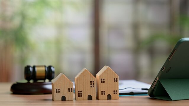 Wooden toy houses stand near judge gavel, digital tablet, document folder placed over office desk surface, representing real estate litigation, property auction, foreclosure law concepts.