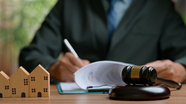 Professional attorney signs real estate agreement papers near wooden gavel, small house models, executing legal property foreclosure auction procedures inside law office workspace, resolving.