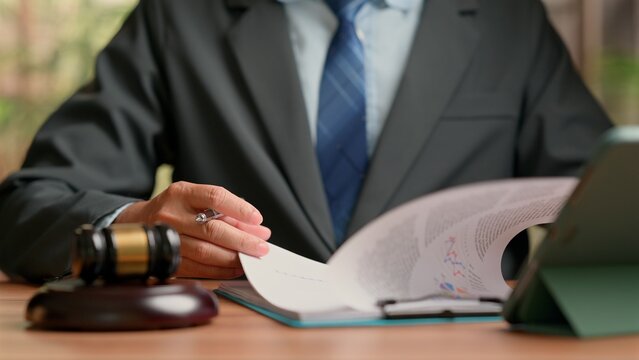Male attorney wearing formal dark suit tie reading paper document holding pen near wooden judge gavel tablet computer sitting inside legal corporate office workspace desk setting.