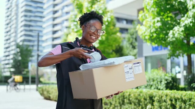 A delivery person walks through a city area while holding a large box. Trees and tall buildings surround the person as they make their way along the path.