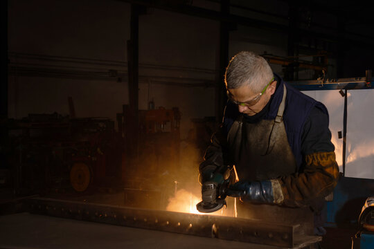 Industrial worker using electric grinder on metal in dark workshop