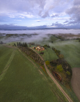 Aerial view of a rustic farmhouse surrounded by cypress trees and rolling green hills blanketed in morning fog in Tuscany, Italy.
