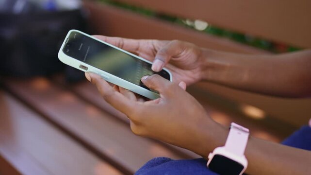 A person sits on a bench in a park and uses a smartphone. They tap the screen while enjoying the outdoors. It is a sunny day with trees in the background.
