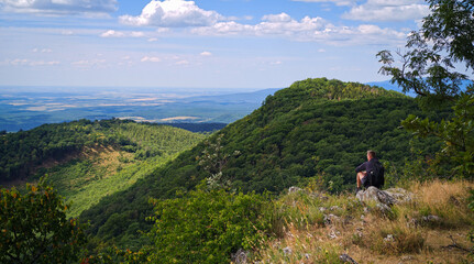 Fototapeta premium Hiker sitting on a rocky viewpoint while enjoying a panoramic landscape of the Bükk Mountains and surrounding forests from Őr-kő, Bükk National Park, Hungary.