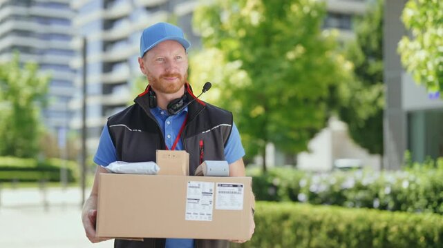 A man stands outside in a city, holding a large cardboard box. He is wearing a blue cap and a black vest. He seems happy while waiting to deliver the package.
