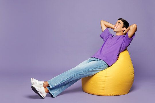 Full body minded smiling happy young man he wear violet t-shirt casual clothes sit in bag chair hold hands behind neck head isolated on plain pastel light purple background studio. Lifestyle concept.