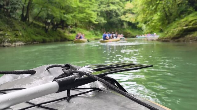 First person view of rafting on a mountain river with a paddle resting on the bow of an inflatable boat, showing a group of people enjoying a water adventure
