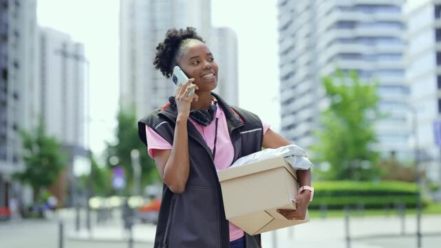 A delivery person stands in a busy city holding a package. She is speaking on her phone while surrounded by tall buildings and greenery. The day appears bright and sunny.