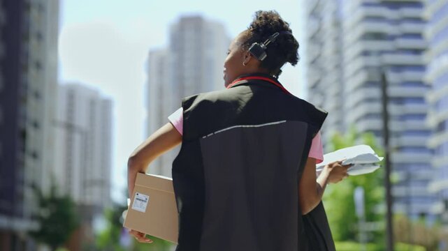 A worker walks along a city street holding a package. Tall buildings are seen in the background as the person heads to a delivery location. The sun is shining and it looks like a busy day.