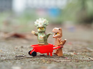 Naklejka premium Indonesia - April 27, 2025 : Mini toy of action figure at outdoor with blurred background. Toy photography concept design. T-rex and triceratops dino characters with trolley wheelbarrow.