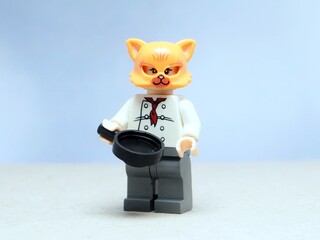 Fototapeta premium Indonesia - May 16, 2025 : Mini toy of action figure at table with blurred background. Miniature photography concept design. Minifigure of orange cat character wearing chef costume with pan and fish.
