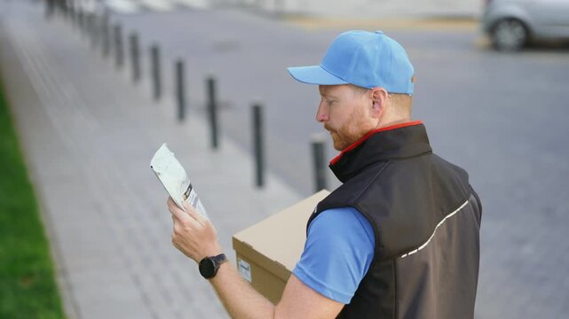 A delivery worker stands on a sidewalk holding packages. He looks up while checking his surroundings in a busy street setting. It appears to be daytime.