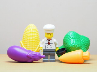 Fototapeta premium Indonesia - February 10, 2026 : Mini toy of action figure at outdoor with blur background. Miniature photography concept design. Minifigure of chef character with eggplant, corn, broccoli and carrot.