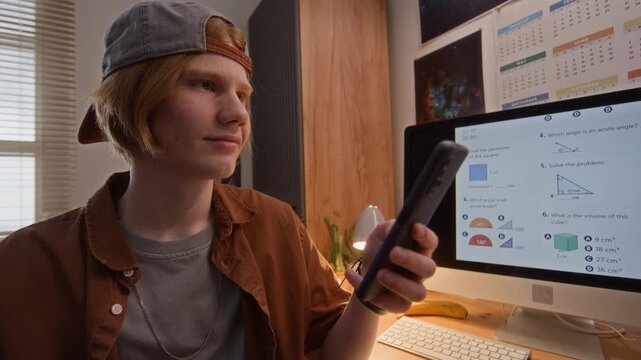 Caucasian teenager holding smartphone while sitting in front of computer monitor displaying math test. Clip is ideal for online education, e-learning, and student distraction concepts