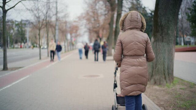 Woman with winter coat pushing baby stroller on city sidewalk during cold autumn day outdoors motion.
