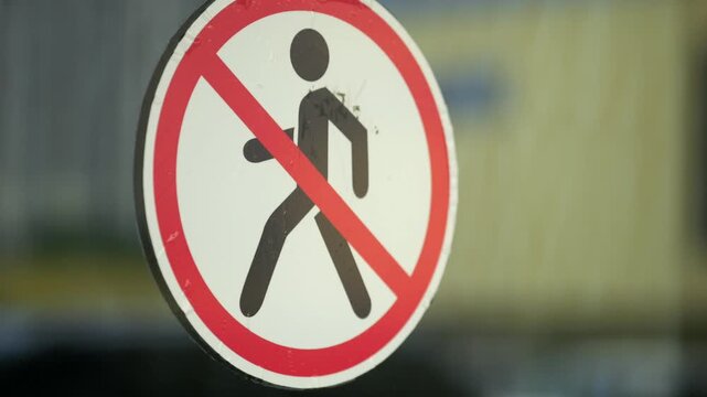 Red circular sign prohibits pedestrians. It features a black pedestrian symbol crossed out. Mounted on a glass surface with reflections. Blurred urban buildings appear in the background