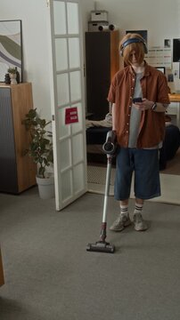 Vertical video of Caucasian teenager using cordless vacuum cleaner while looking at smartphone. Footage is perfect for smart home appliance ads or youth lifestyle campaigns