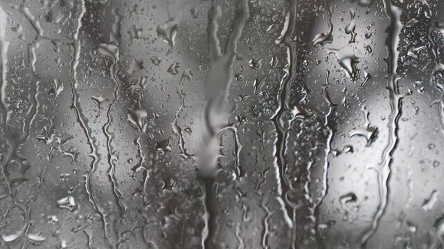A detailed macro shot of heavy raindrops and water droplets streaming down a glass pane during a rainy day. The abstract interplay of light, moisture, and flowing water creates a moody, textured