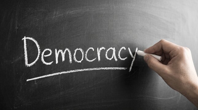 A human hand writes the word "Democracy" in white chalk on a dark blackboard, emphasizing governance and political concepts.