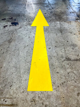 Yellow directional arrow painted on a basement parking floor, guiding drivers along designated routes and improving vehicle navigation, circulation, and safety within the parking facility.