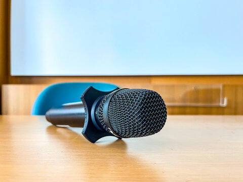 Close-up of a black handheld microphone resting on a wooden classroom table, ready for presentations, lectures, seminars, meetings, and public speaking events.