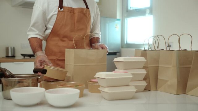Male chef packaging to paper bags freshly prepared fresh meals for online food delivery in a small business kitchen. Cloud kitchen, local restaurant, food startup. Meal prep and entrepreneur concept.
