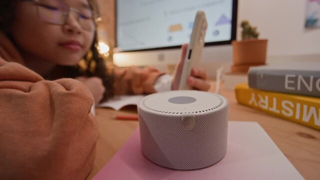 Asian teen girl with glasses using smartphone and smart speaker while studying geometry at desk. Ideal for marketing campaigns promoting smart home devices, e-learning platforms, and edtech
