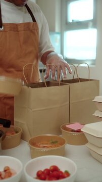 Male chef packaging to paper bags freshly prepared fresh meals for online food delivery in a small business kitchen. Cloud kitchen, local restaurant, food startup. Meal prep and entrepreneur concept.
