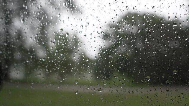Close up macro view of rain drops sliding and trickling down a glass window pane on a rainy day, with a soft blurred background creating a calm and moody atmosphere.