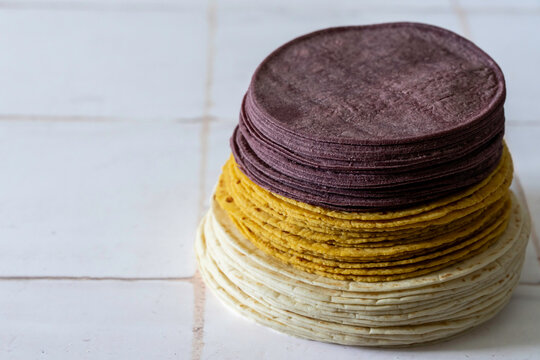 Blue corn, flour and corn and flour tortilla shells stacked on a white tile surface 