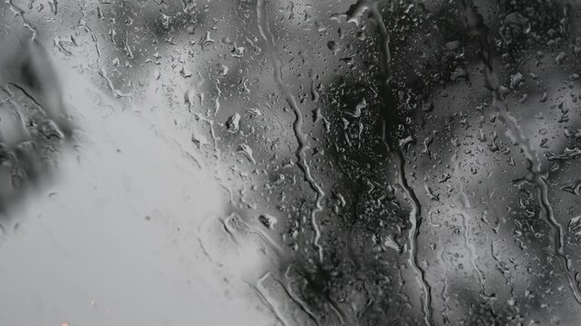 Close up macro view of rain drops sliding and trickling down a glass window pane on a rainy day, with a soft blurred background creating a calm and moody atmosphere.