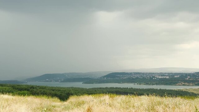 A heavy summer shower pours over a scenic reservoir and forests in Simferopol, Crimea. Dry grassy hills wait under the dark sky, creating a peaceful and moody feeling of awe.