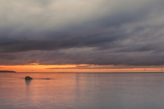 Sunset over calm ocean with dark clouds and rock in water