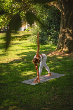 Young woman in white activewear holding the triangle yoga pose with one arm reaching to the sky on a mat in a green park a grounding stretch for strength and balance.