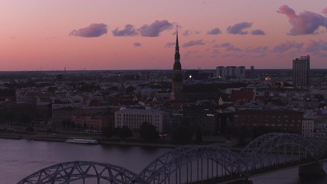 Aerial Riga at sunset shows St. Peter's Church spire, Dzelzcela Railway Bridge, Daugava River, old town roofs, modern skyline, boat gliding, clouds drifting.